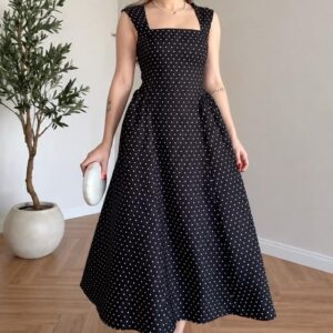 Femme portant une robe longue noire à pois blancs avec col carré, larges bretelles et jupe fluide évasée.